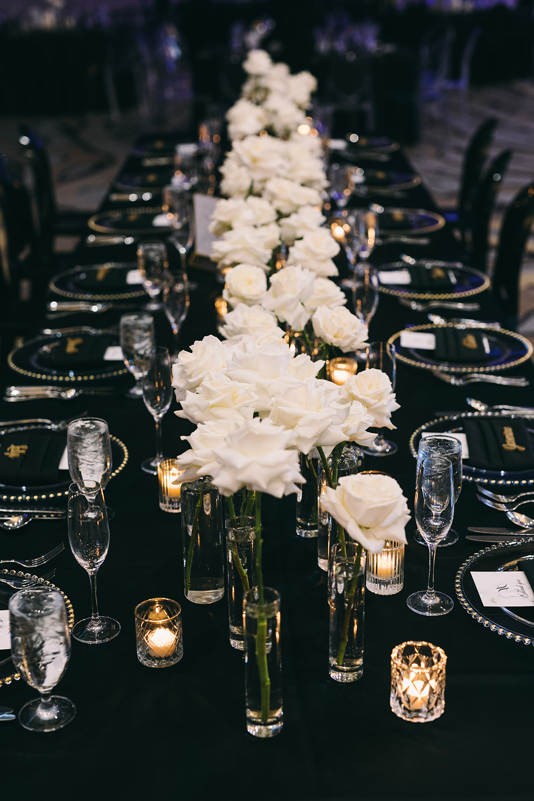 Long table with black cloth and white roses as centerpieces and black silverware with tiny votive candles