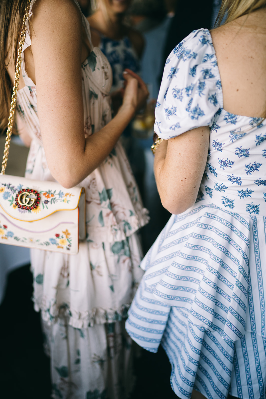 Two women standing. One of them is wearing a blue and white dress and the other is wearing a flowery dress holding a Gucci bag
