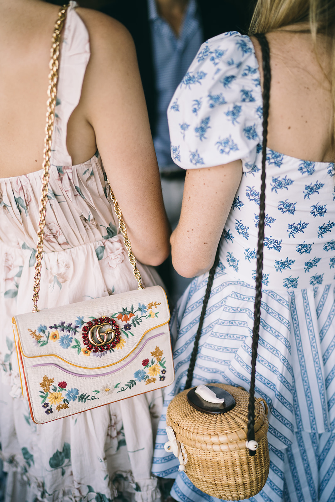 Two woman standing with their backs to camera one has a wicker purse and the other has a purse with flowers on it 