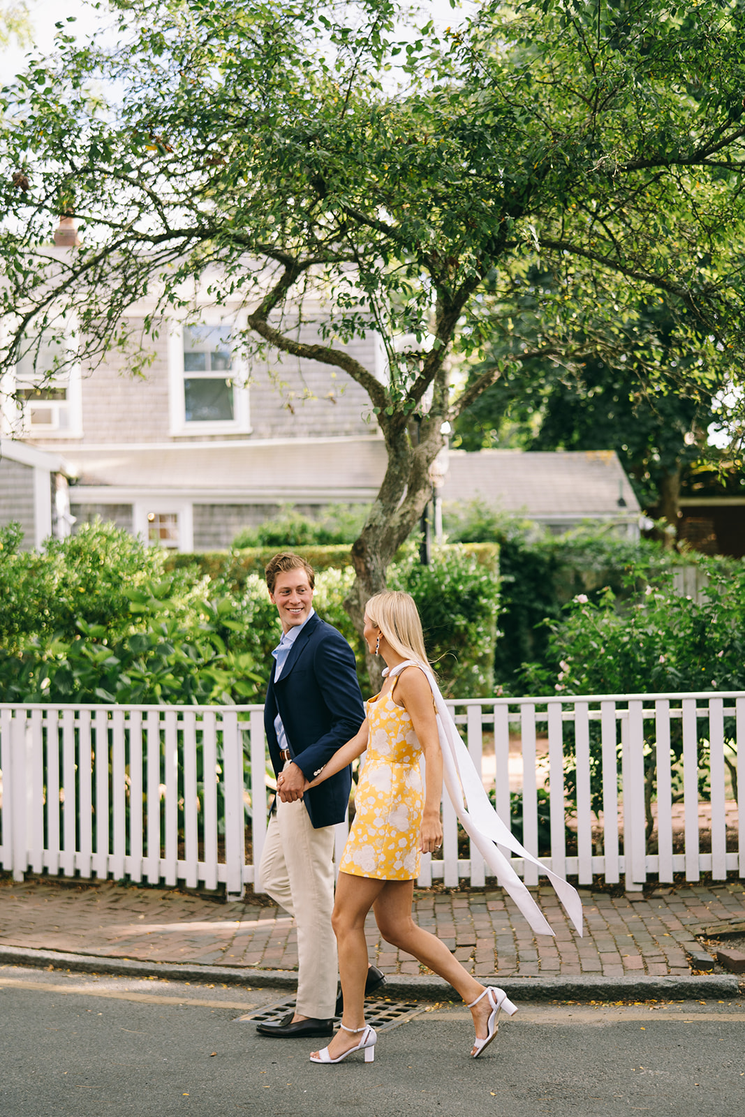 Engaged couple looking at each other smiling as they walk down neighborhood street with trees in the background