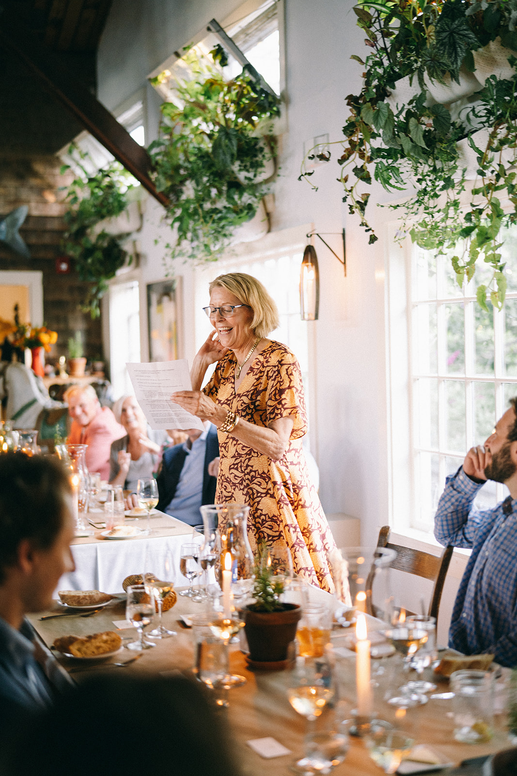 Woman in flowery dress standing up holding a piece of paper and speaking to everyone else in the room