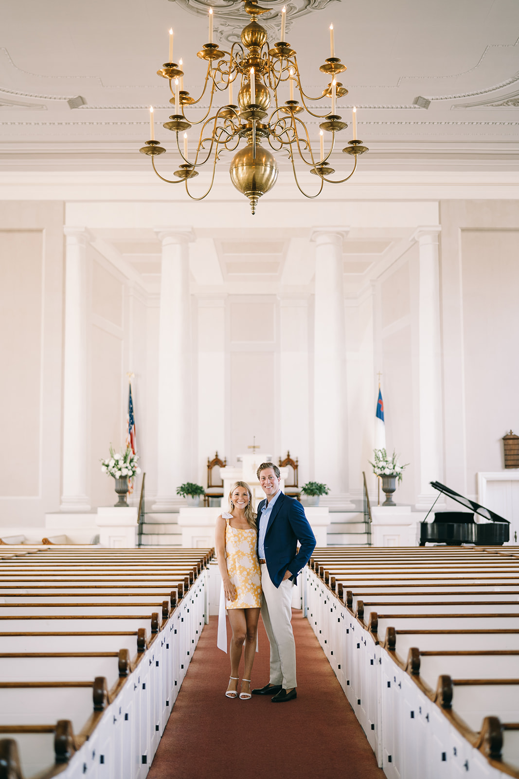 Couple standing together smiling in a church with high ceiling and white walls and a golden chandelier at a church in Nantucket