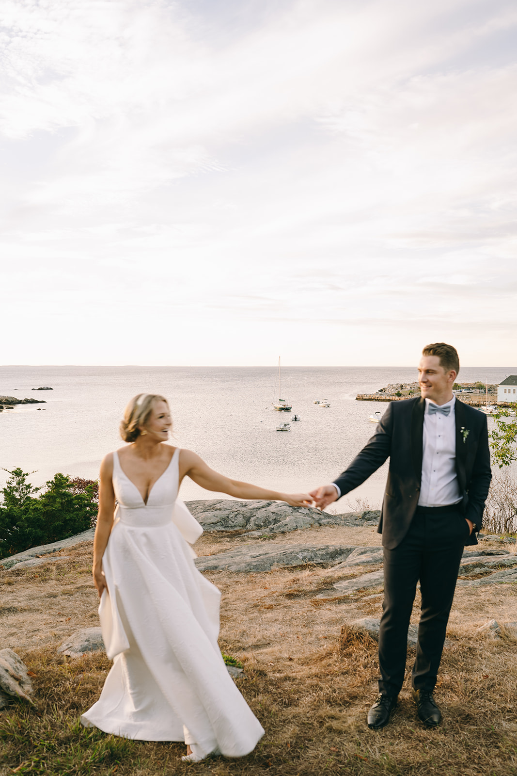 Bride and groom leaning apart but holding each other's hand looking at each other with the bay in the background
